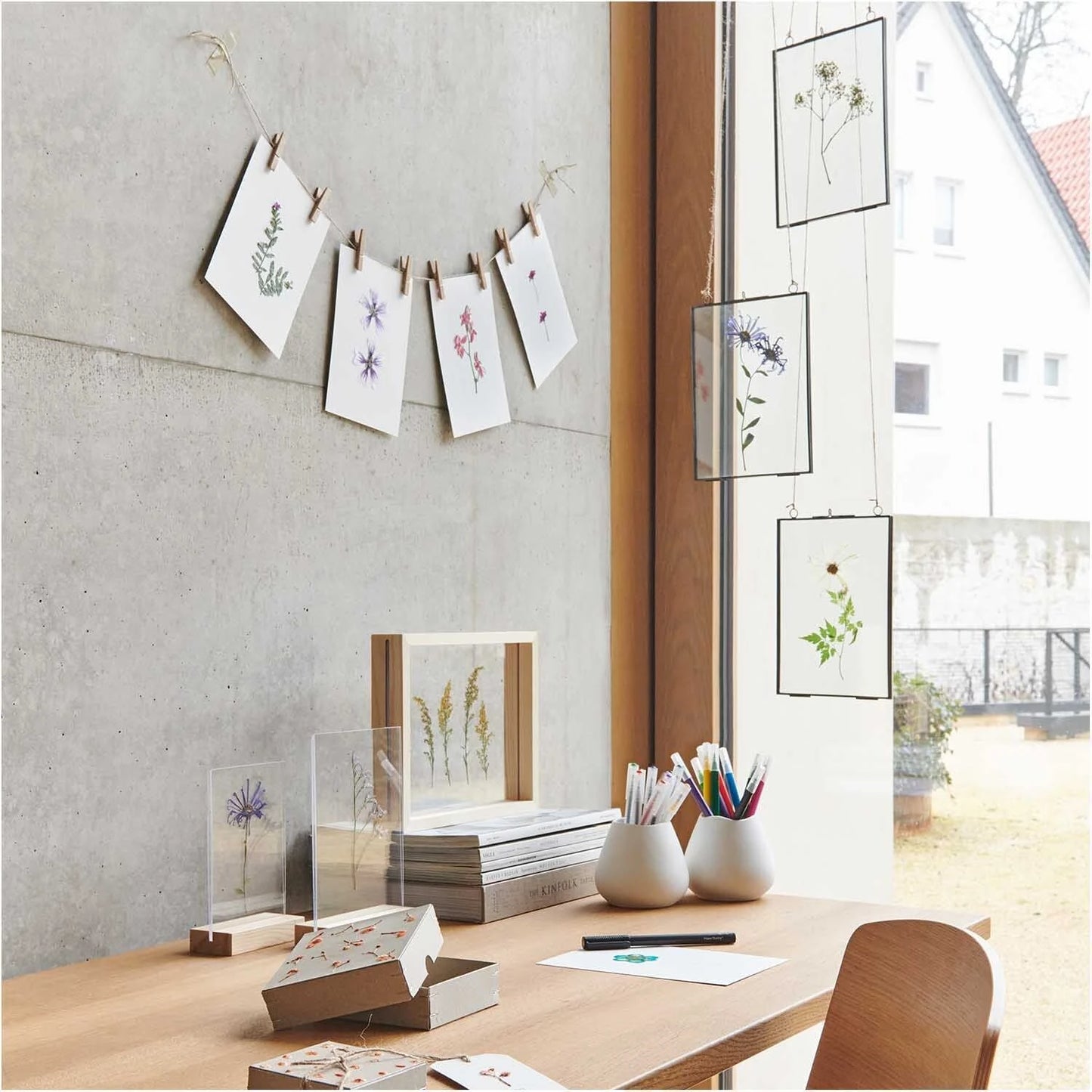 una habitación con una mesa donde hay unos soportes con base de madera y dos paneles acrílicos transparentes con flores dentro que hacen efecto como si flotara, un par de portalápices marcos transparentes colgados de la ventana con flores dentro