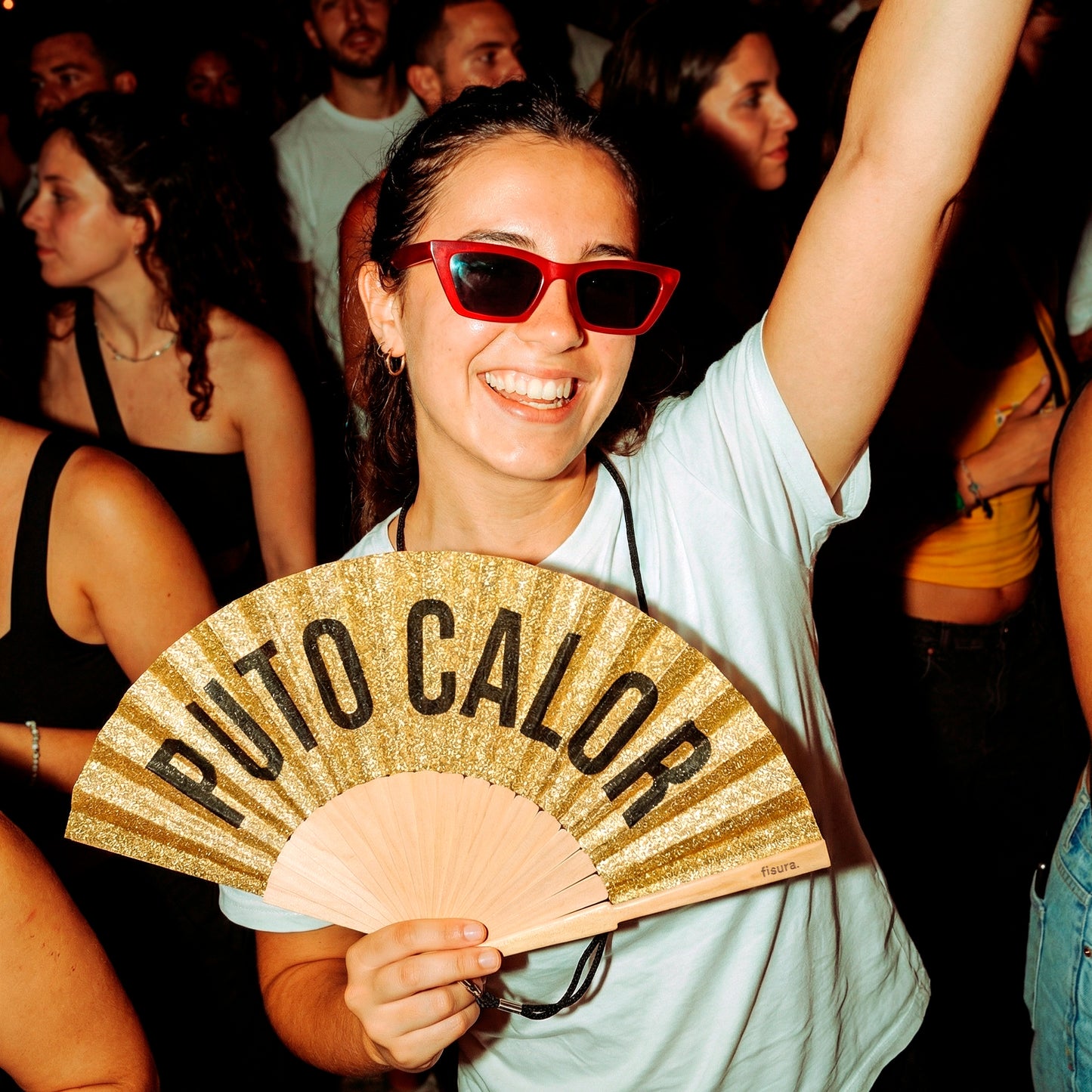 chica con el abanico de la marca fisura con purpurina dorada y el texto "PUTO CALOR" en color negro en una verbena de noche