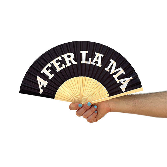 Abanico negro con A fer la mà escrito en blanco, sostenido por una mano sobre un fondo blanco.