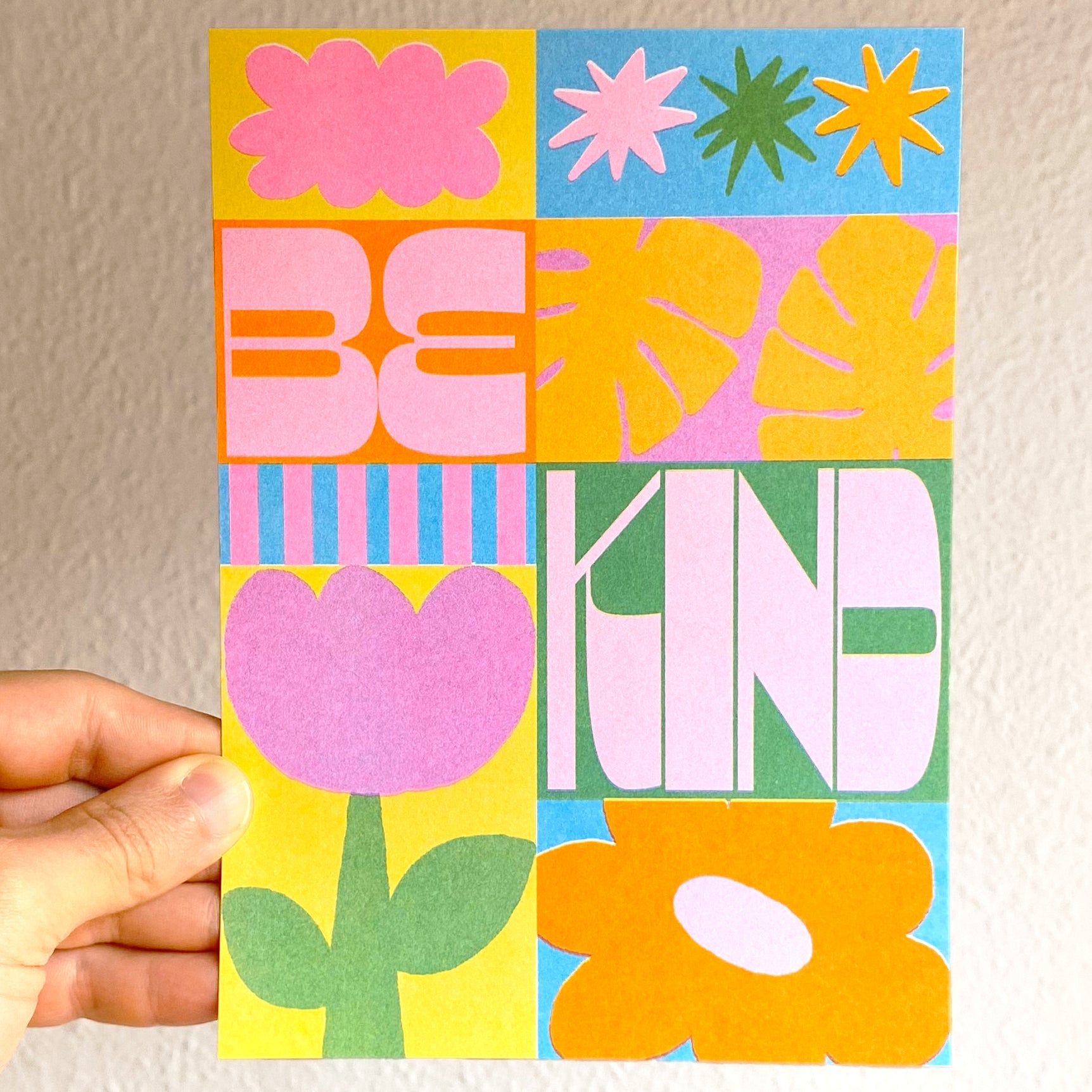 Mano sujetando la mini print de colores con flores, hojas, estrellas y el mensaje BE KIND