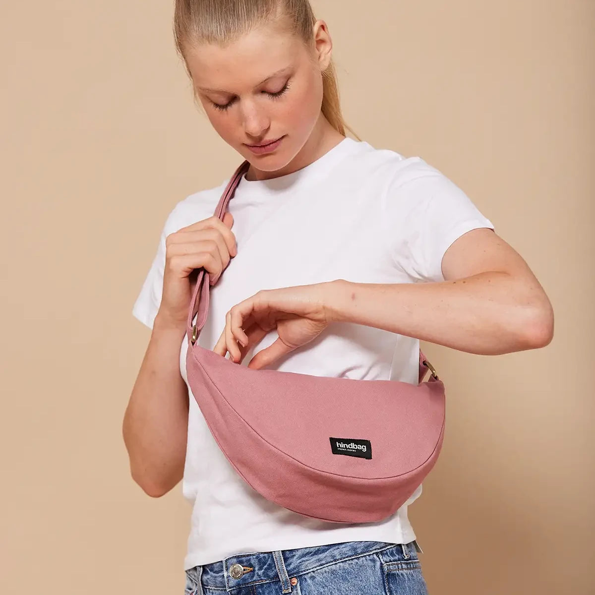 Mujer con camiseta blanca abriendo el bolso riñonera rosa de la marca Hindbag con forma de media luna