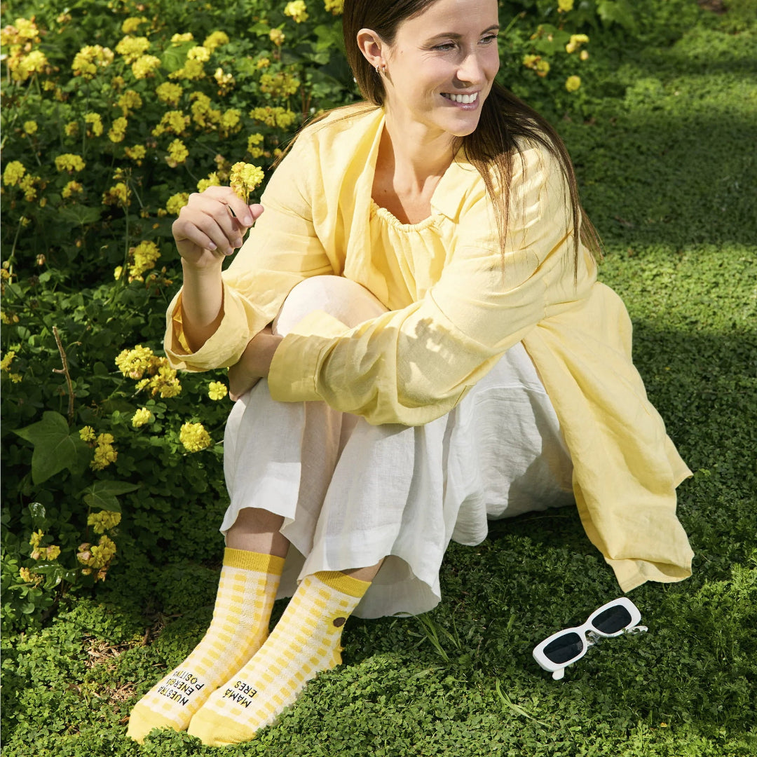 mujer sentada en la hierba con camisa amarilla y los calcetines de caña baja estampados a cuadros amarillos con el mensaje "Mamá eres nuestra energía positiva"
