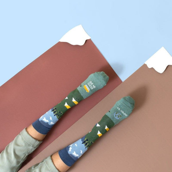 Pies con los calcetines con estampado de montañas tiendas de campaña, una camper y el globo del mundo y el mensaje Que te quiten lo viajao sobre montañas de cartulina con nieve en la punta