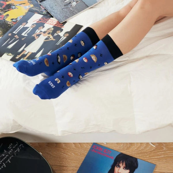 Piernas de mujer en una cama llena de vinilos con los calcetines azules con el mensaje Eres un temazo y estampado de rayos, notas musicales y caras de iconos de la música