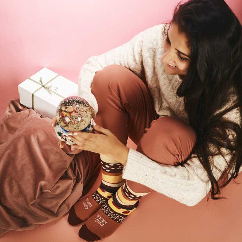 Mujer con bebida de Navidad, regalos y los calcetines de Navidad estampados de capas de chocolate, almendra, fruta escarchada, almendrado, turrón blando y el mensaje "Estás mejor que el turrón"