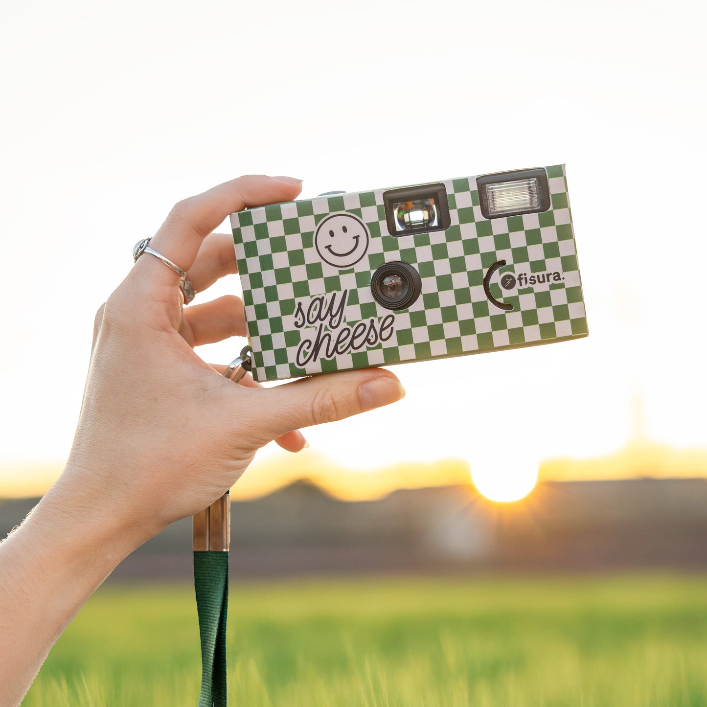 Mujer sosteniendo una cámara desechable con aires retro a cuadros verdes y beiges con correa para colgar en un campo verde con el atardecer de fondo