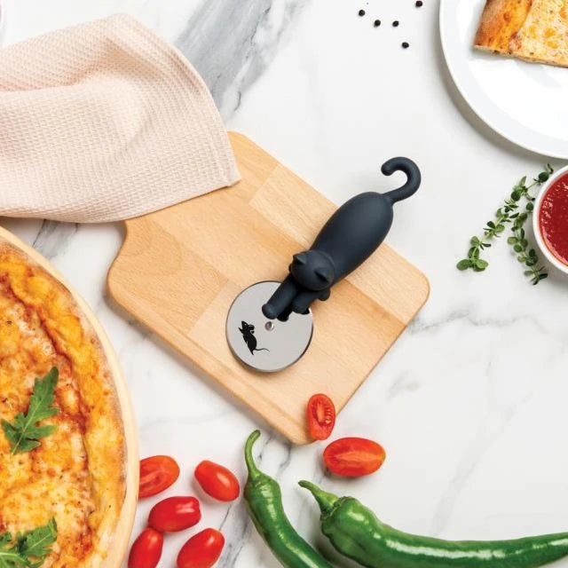 Una mesa de mármol con pimientos, tomates cherry, una pizza, un trapo y un cortapizzas con forma de gato sobre una tabla de madera