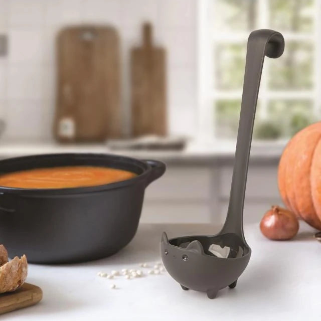 Cucharón de cocina con forma de gato con el rabo levantado junto a una olla con puré de calabaza