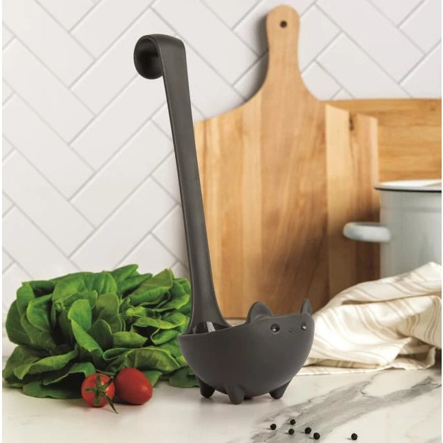 Cucharón de cocina con forma de gato con el rabo levantado en una encimera de ocina con lechugas, tomates y una tabla de cortar