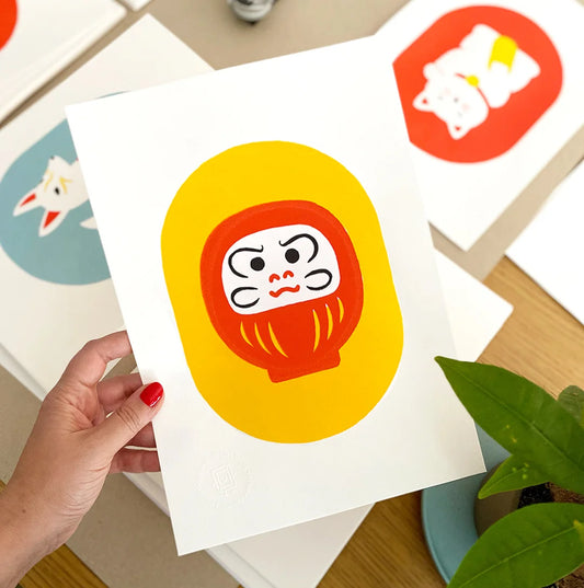 Mano sujetando una print de estilo japonés con un Daruma rojo sobre fondo amarillo. Al fondo se ven otras ilustraciones con motivos japoneses. Una planta aporta un toque natural a la escena.