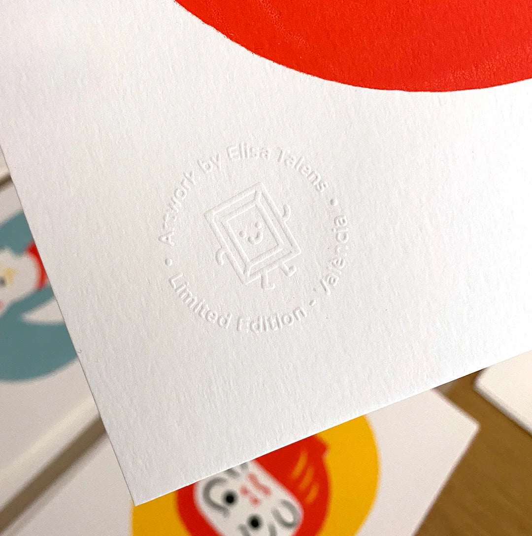 Detalle del cuño seco en relieve sobre el papel de una print. El sello circular dice “Artwork by Elisa Talens · Limited Edition · Valencia” e incluye un dibujo simpático de un marco con patas y carita sonriente. El fondo muestra otras ilustraciones con colores vivos.