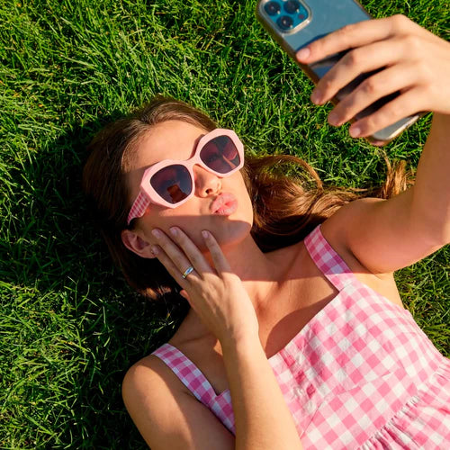 Mujer con vestido de cuadros vichy rosas y gafas de sol de montura rosa chicle haciéndose un selfie tumbada sobre el césped