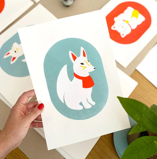 Mano sujetando una print de estilo japonés con un Kitsune blanco con detalles rojos y amarillos sobre fondo azul. Al fondo se ven otras ilustraciones con motivos japoneses. Una planta aporta un toque natural a la escena.