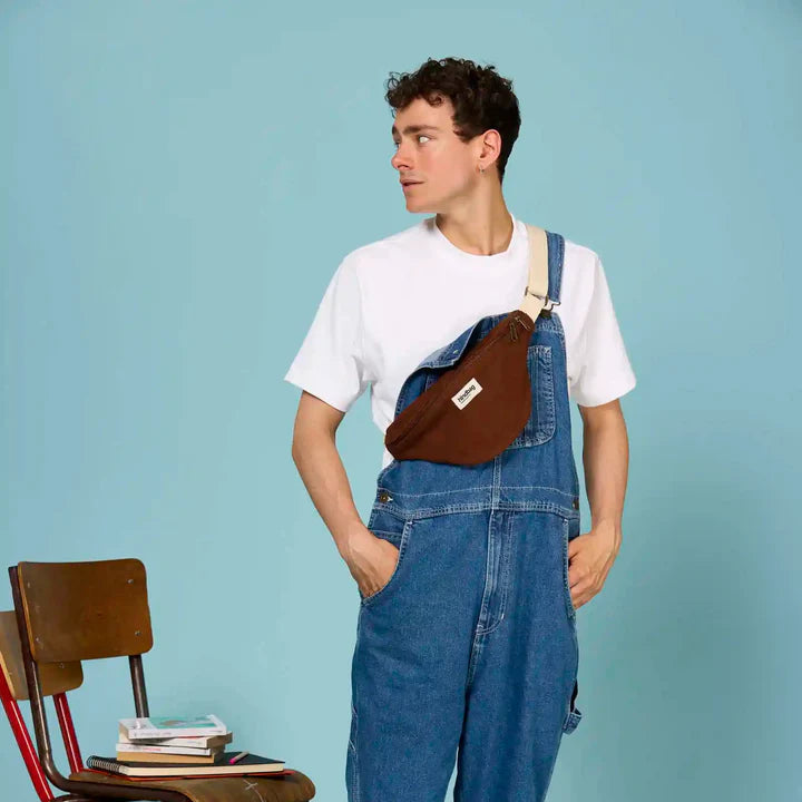 Chico con mono vaquero y camiseta blanca con una riñonera de Hindbag color marrón chocolate