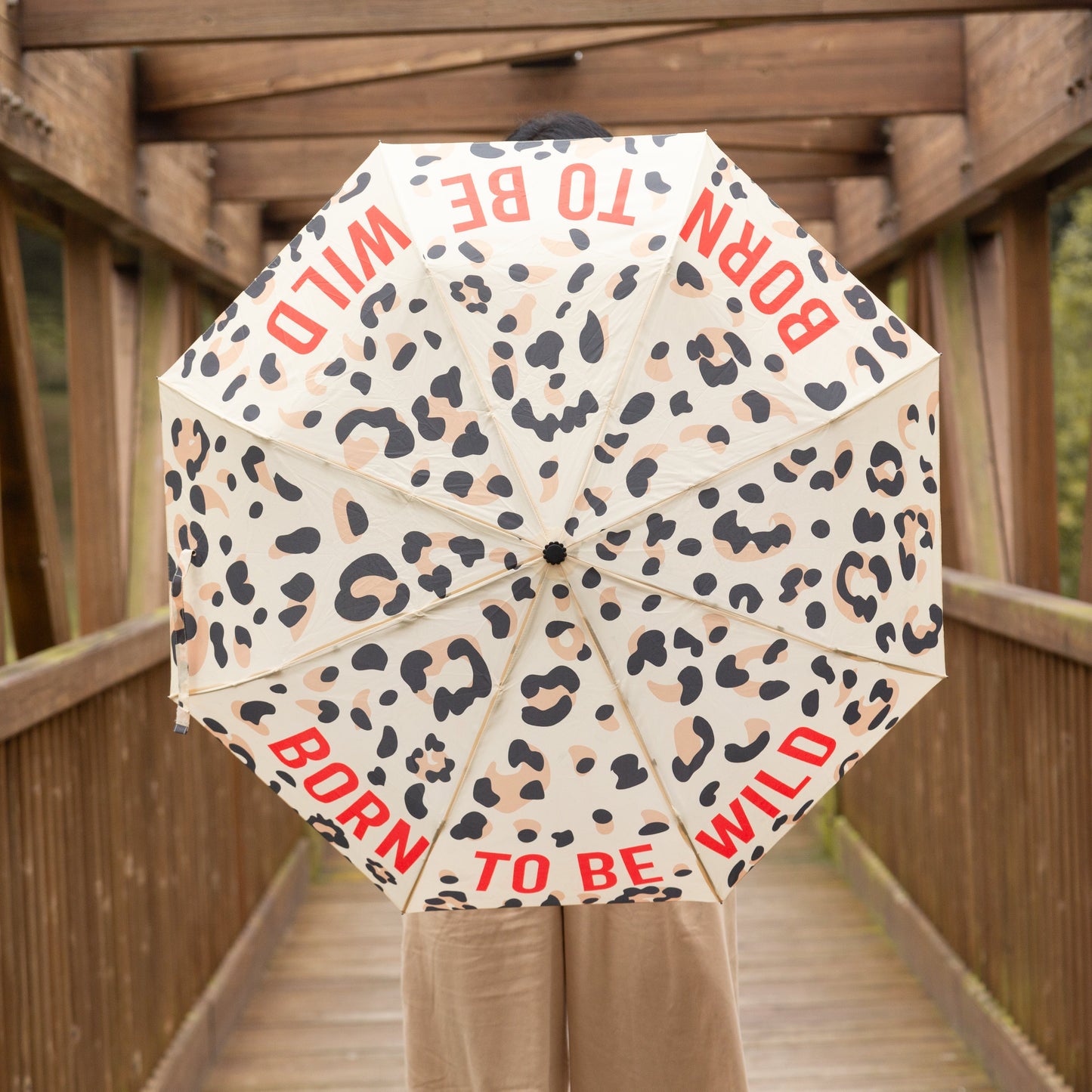 Mujer en un puente de madera con un paraguas plegable con esqueleto de fibra de vidrio negro, estampado de leopardo y el mensaje Born to be wild escrito en letras mayúsculas rojas