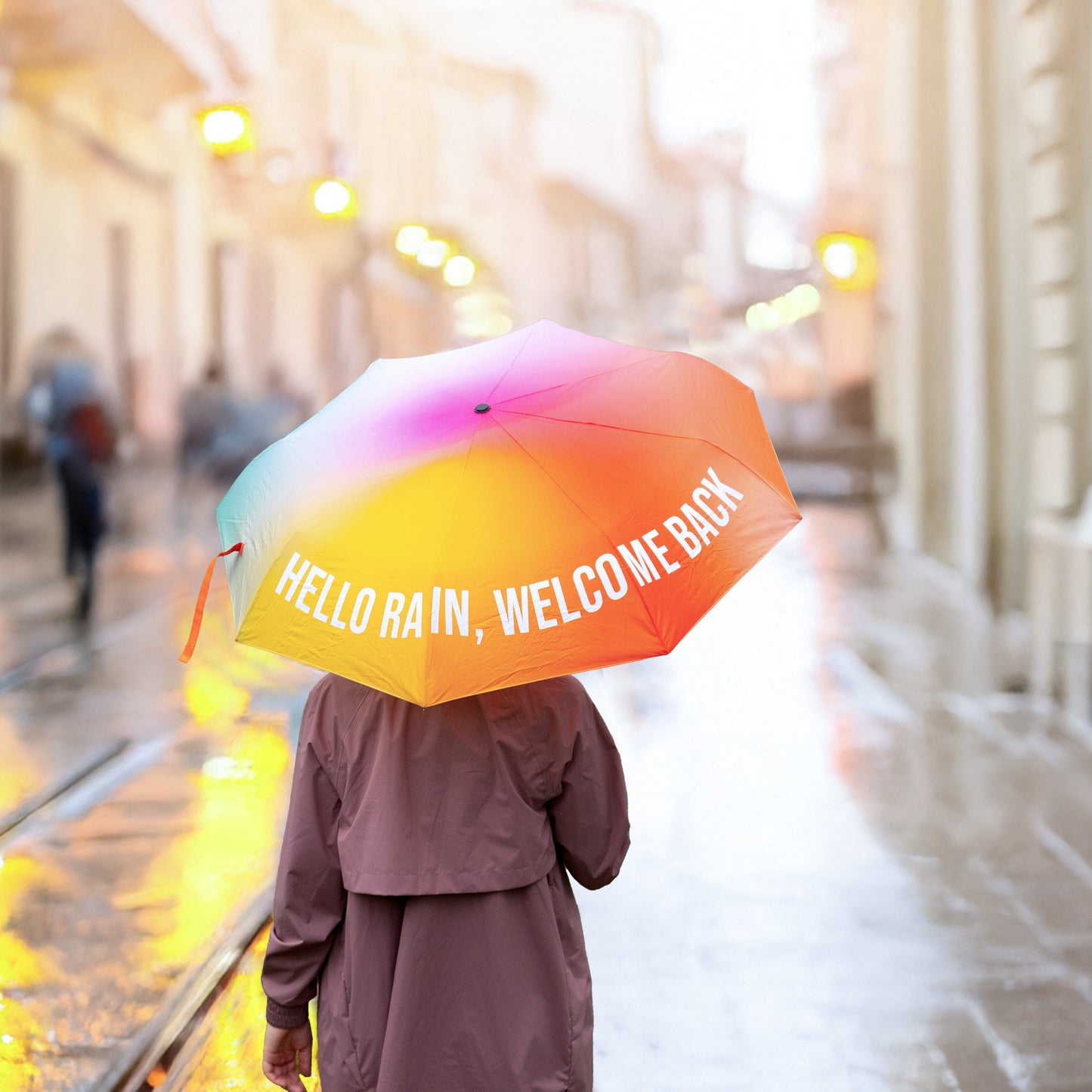 Chica con chubasquero andando por la calle con un paraguas plegable estampado con un degradado de colores y el mensaje Hello rain, welcome back en letras mayúsculas blancas