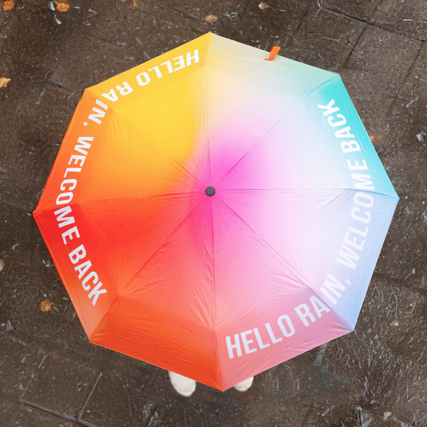 Paraguas plegable con estampado degradado de colores y el mensaje Hello rain, welcome back en letras mayúsculas blancas visto desde arriba