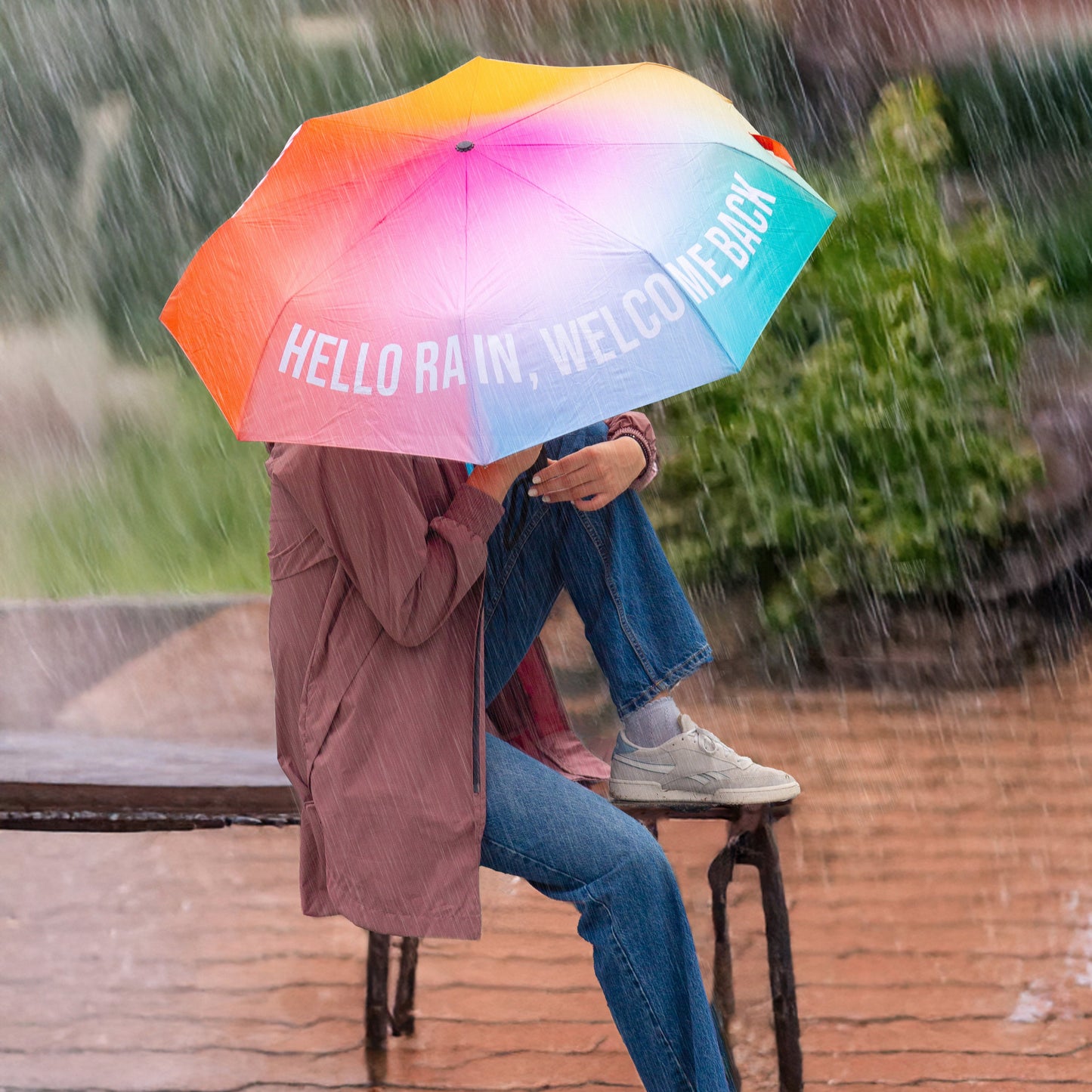 Chica bajo la lluvia con un paraguas plegable con esqueleto negro y estampado degradado de colores con el mensaje Hello rain, welcome back en letras mayúsculas blancas