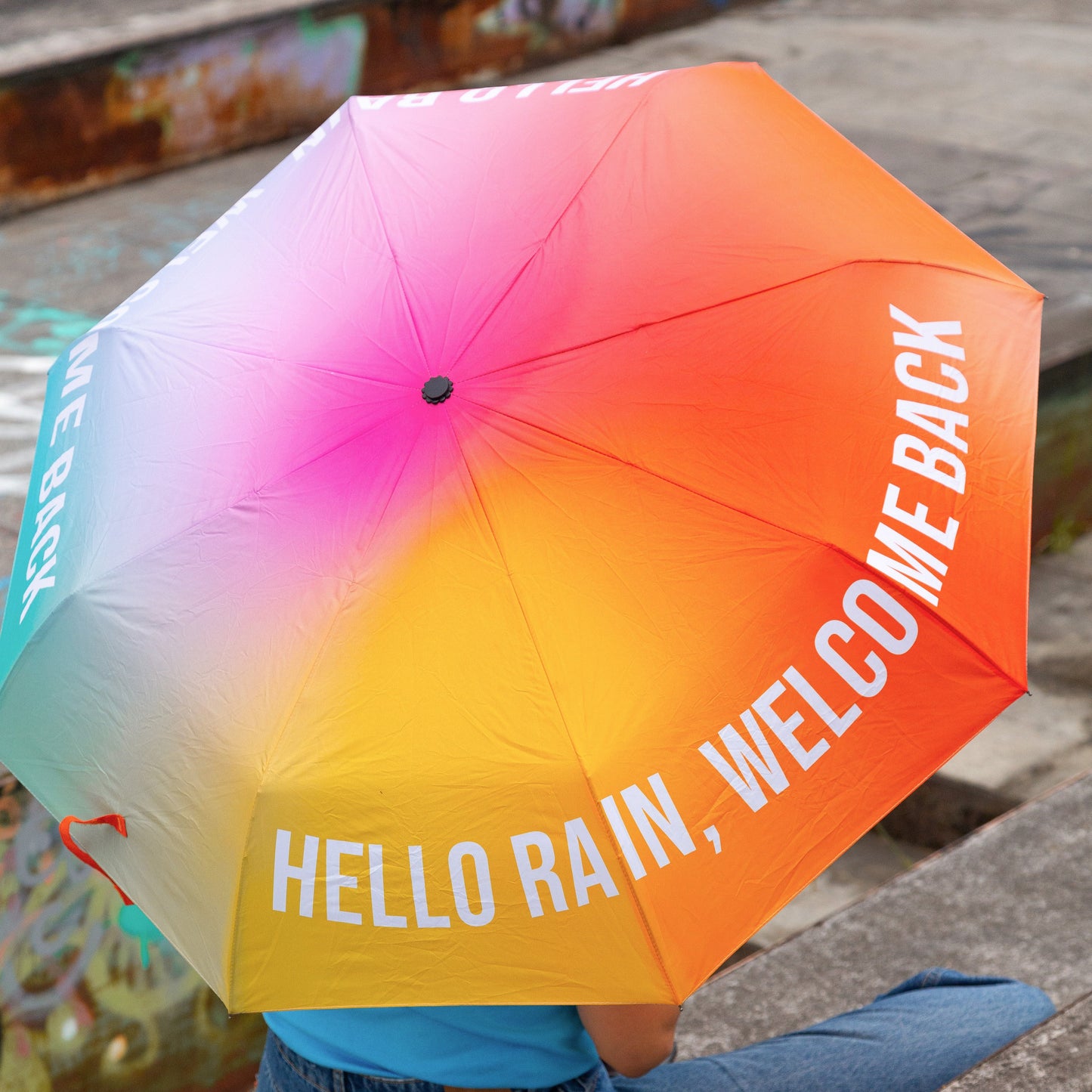Chica sentada de espaldas con el paraguas plegable con esqueleto negro y estampado degradado de colores con el mensaje Hello rain, welcome back en letras mayúsculas blancas