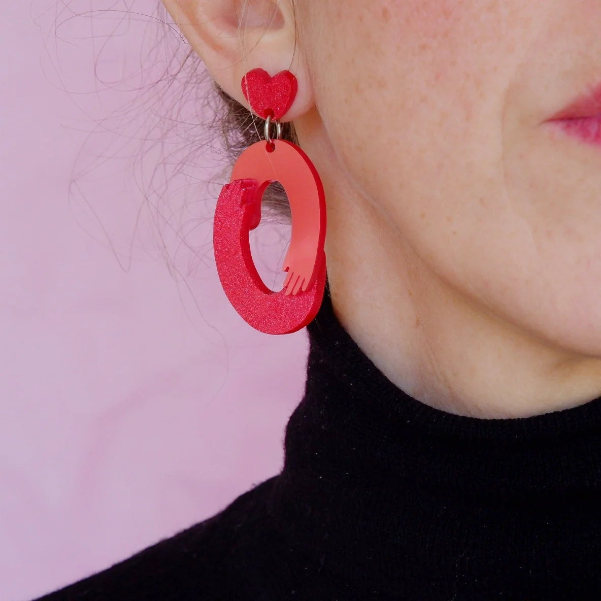 Primer plano de una persona usando un pendiente en tonos rojos con forma de óvalo y brazos que simulan un abrazo. La pieza está unida a un pequeño corazón en la parte superior.
