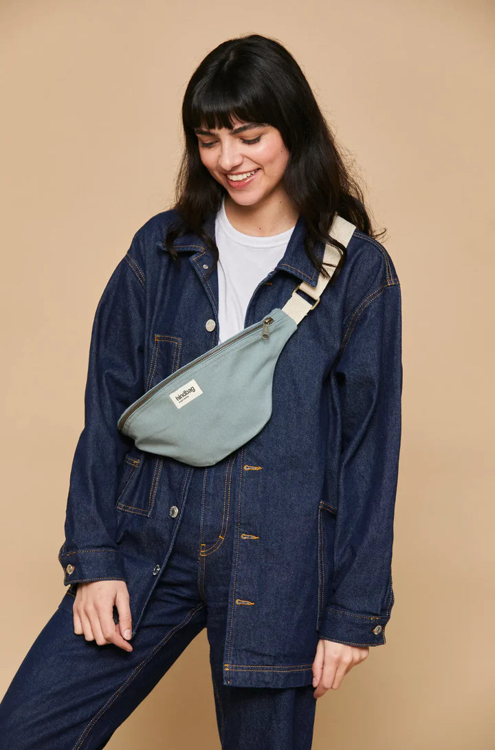 Mujer joven vestida de vaquero con la riñonera de la marca francesa Hindbag color azul verdoso salvia y correa blanca