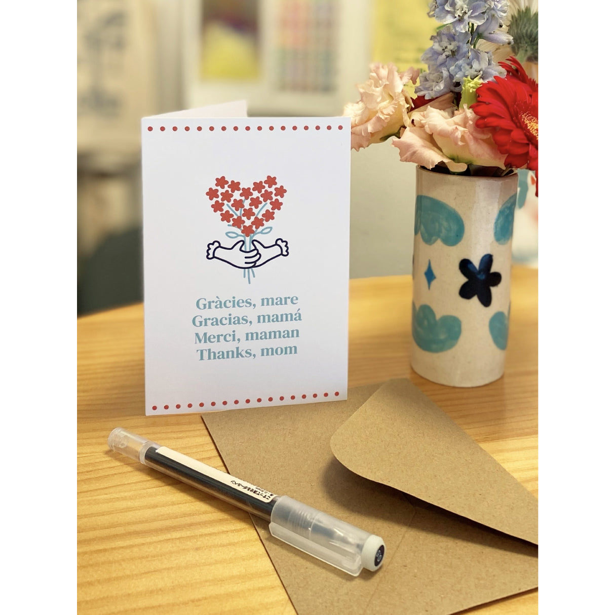 Fotografía de una tarjeta de felicitación sobre una mesa de madera, junto a un bolígrafo, un sobre kraft y un jarrón con flores. La tarjeta muestra un ramo de flores rojas en forma de corazón, sujetado por unas manos, con el mensaje "Gràcies, mare. Gracias, mamá. Merci, maman. Thanks, mom".