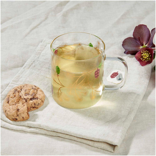 Taza de té cristal con adornos navideños y florales en dorado sobre un paño y junto a una galletita