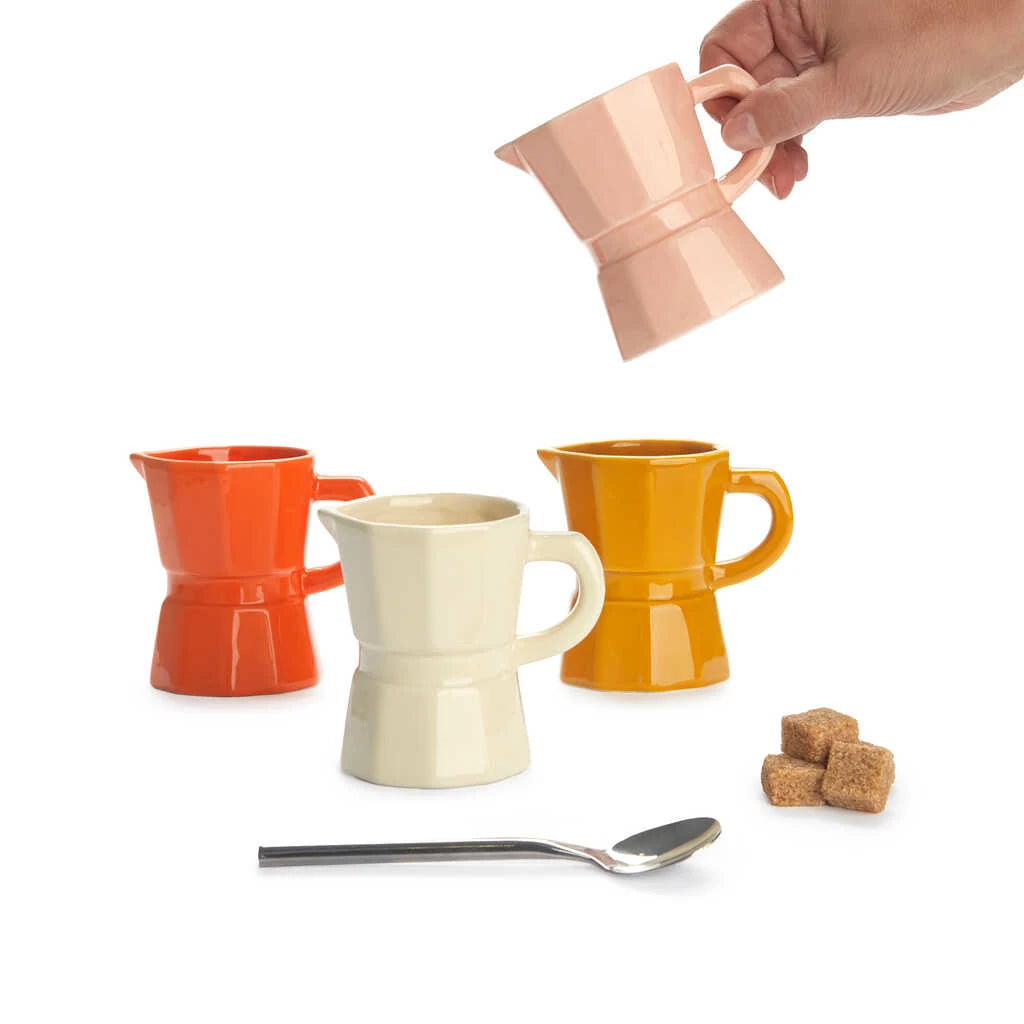 Mano sosteniendo una de las tazas del juego de cuatro tazas de cerámica con forma de cafetera italiana para café cortado en colores pastel: naranja, crema, rosa y mostaza junto a una cucharilla y un terrón de azúcar