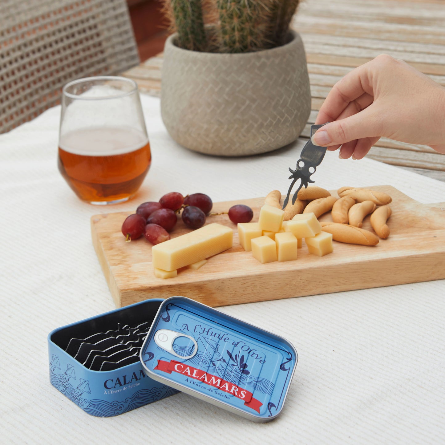 Mesa con tabla de quesos y uvas y una cerveza y alguien pinchando con un tenedor de aperitivo con forma de calamar pequeñito