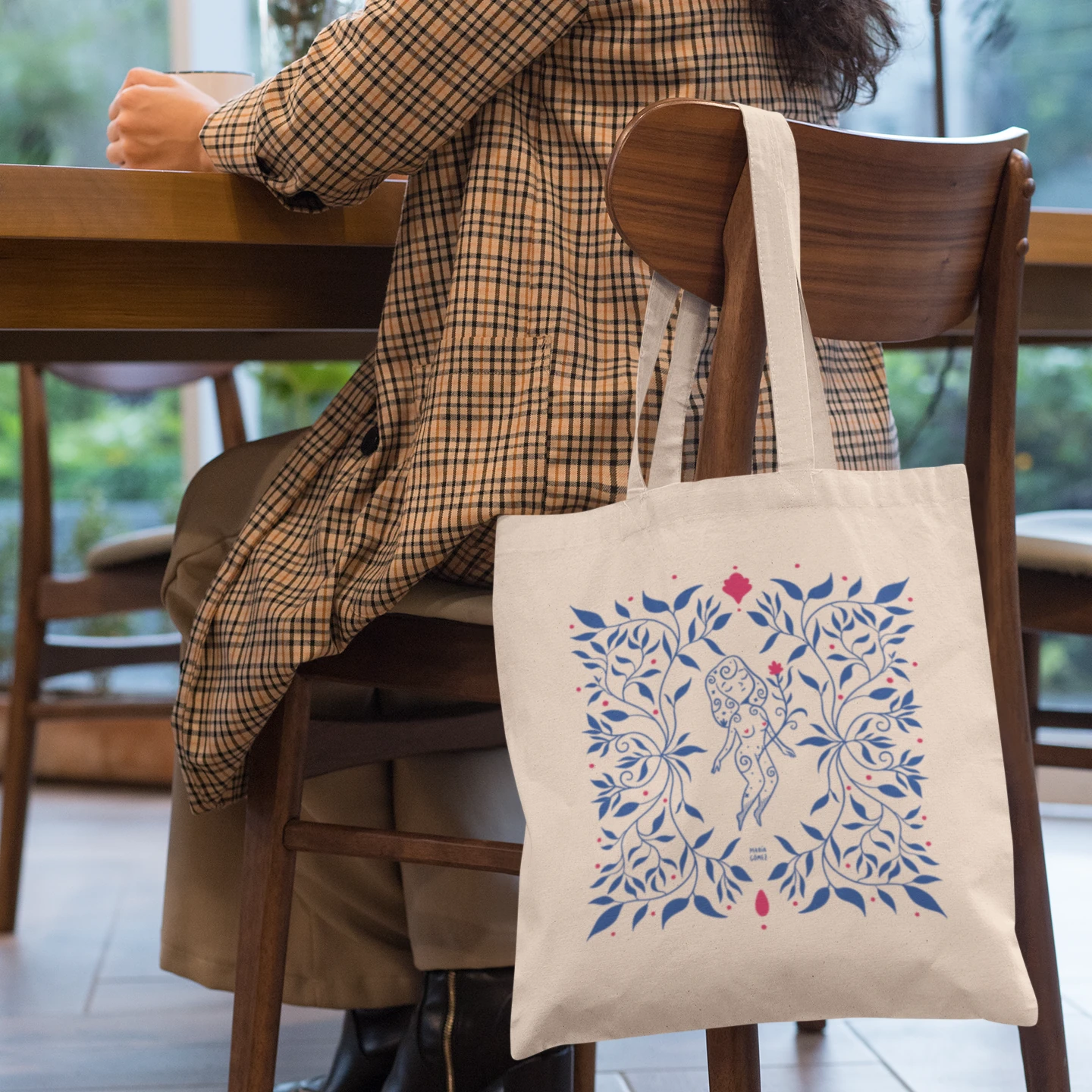 Mujer en una silla de madera tomando café con la bolsa de tela con una mujer flotando entre flores azules de María Gómez