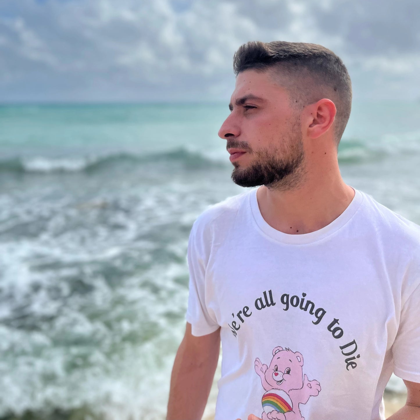 Joven de perfil en la playa con la camiseta blanca con un oso amoroso que dice We're all going to die