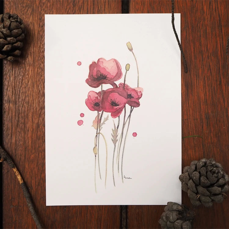 Lámina con unas amapolas pintadas en acuarela de la ilustradora Marialu sobre una mesa de madera con adornos de piñas