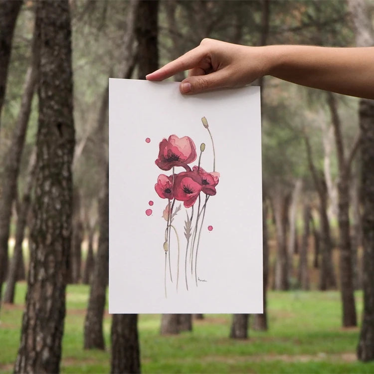 Lámina con unas amapolas pintadas en acuarela de la ilustradora Marialu sobre un fondo de un bosque con una mano sujetándola
