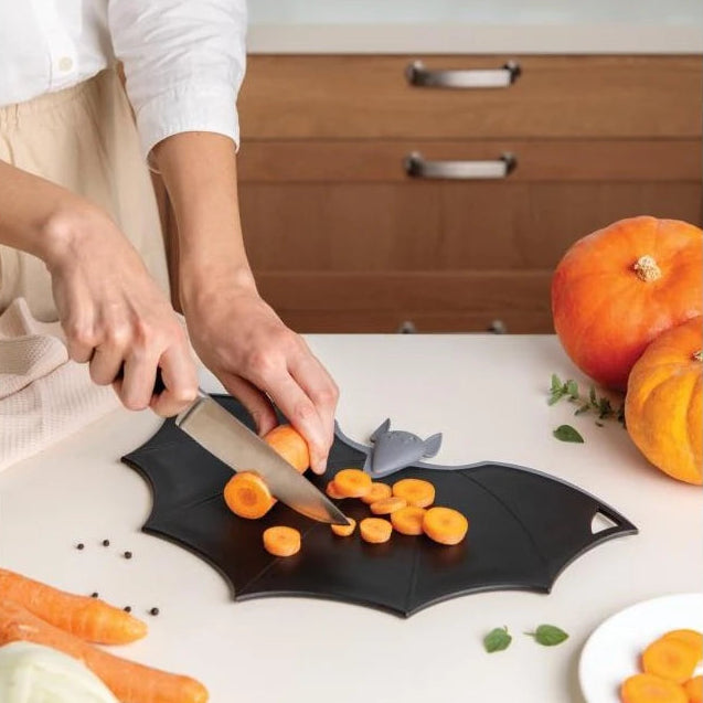 Persona cortando zanahorias sobre una tabla de cortar plegable con forma de murciélago con calabazas encima de la mesa de la cocina