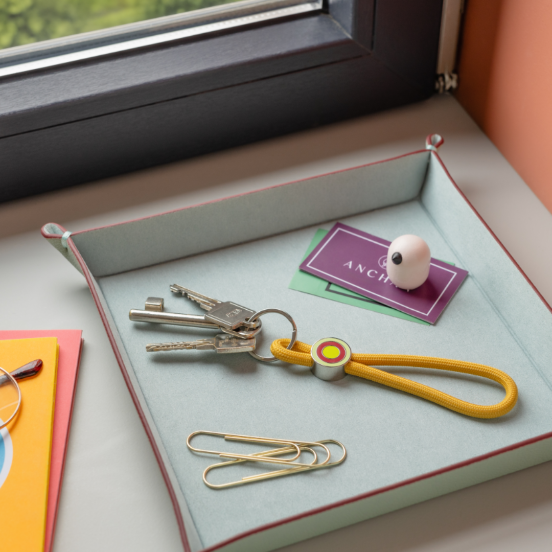 llavero colgante de cuerda amarillo mostaza para el móvil o las llaves con anilla plateada y un enganche con círculos de colores con llaves enganchadas y dejada en una caja con unos clips
