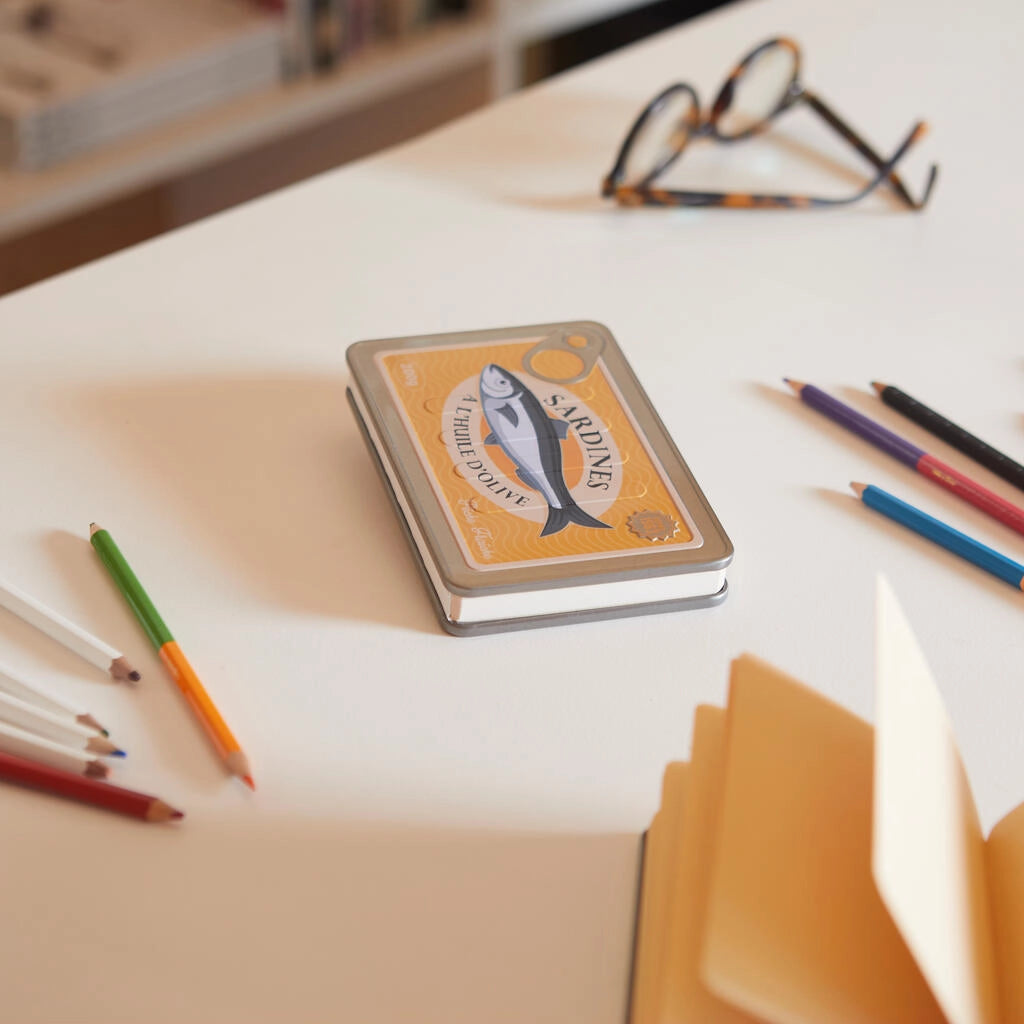 libreta en forma de lata de conservas en color amarillo y con partes metálicas y una sardina en el centro con el texto Sardines a l'huile d'olive, cerrada sobre una mesa con bolis y unas gafas