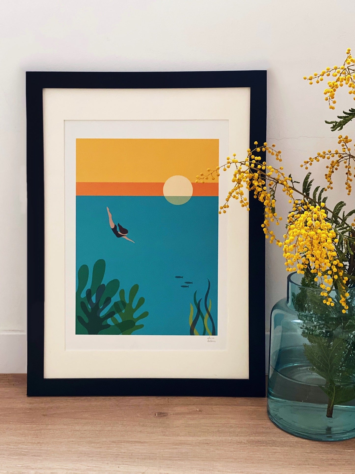 Un marco en color negro para fotos o prints con una lámina de Elisa Talens en la que se ve na mujer buceando en el mar está apoyado sobre una pared junto a un jarrón con flores amarillas.