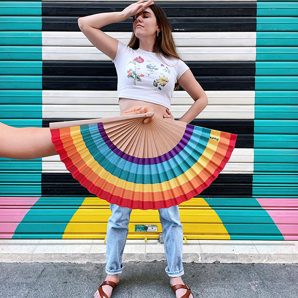 Mano sujetando un abanico con los colores del arcoiris y una mujer posando con orgullo de fondo