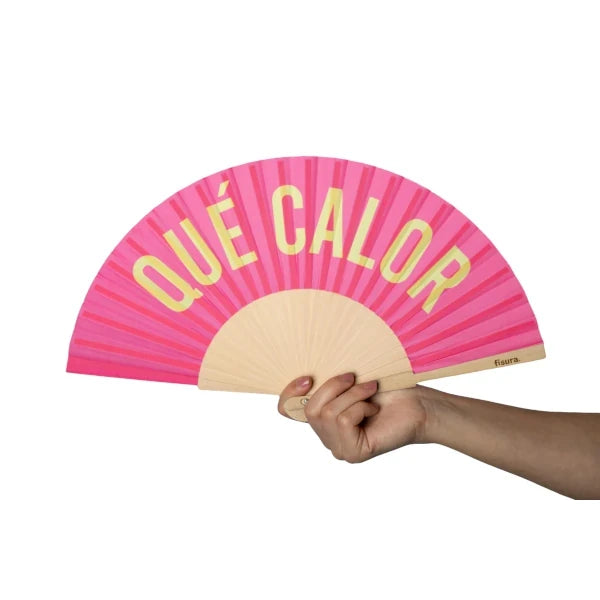 Mano sosteniendo el abanico rosa chicle de tela y madera con el mensaje QUÉ CALOR en letras mayúsculas amarillas