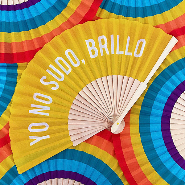 Abanico amarillo con el mensaje "Yo no sudo, brillo" en letras mayúsculas blancas y varillas de madera sobre un montón de abanicos arcoiris con los colores del orgullo