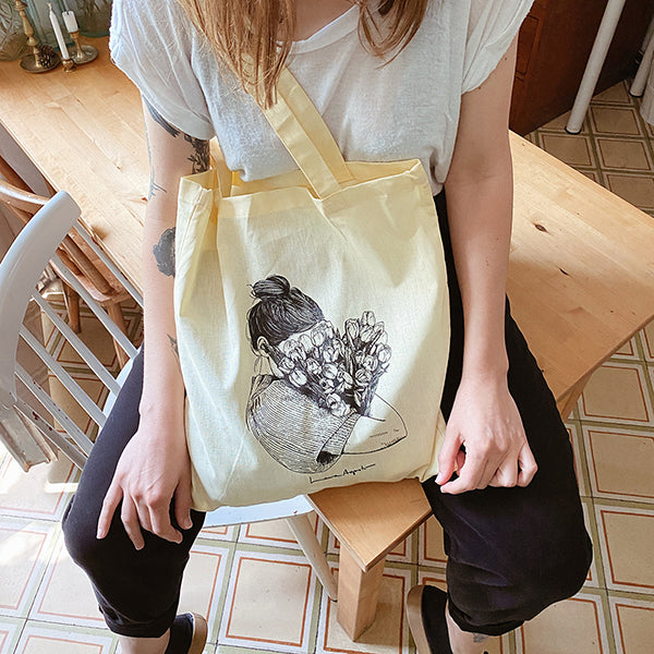 Chica sentada en una mesa de madera con la totebag con una ilustración de Laura Agustí serigrafiada de una mujer con el peli recogido hundiendo la cabeza en un ramo de tulipanes