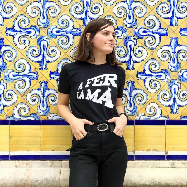 Mujer con camiseta negra en la que se lee la expresión típica de la Comunidad Valenciana A fer la mà en letras blancas apoyada sobre una pared de azulejos azules y amarillos en Valencia