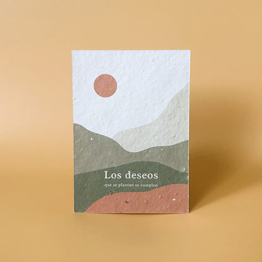 Tarjeta con semillas de Sheedo con un paisaje en tonos verdes y tierra con un sol naranja y el mensaje "Los deseos que se plantan se cumplen"