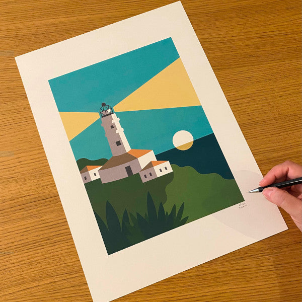 Ilustración tamaño A3 de Elisa Talens con un faro encendido al atardecer, con el sol poniéndose en el mar de fondo y casitas y vegetación en primer plano sobre una mesa de madera