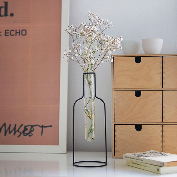 Florero con forma de silueta de botella de hierro con una probeta de cristal que contiene un tallo de flores secas encima de un escritorio