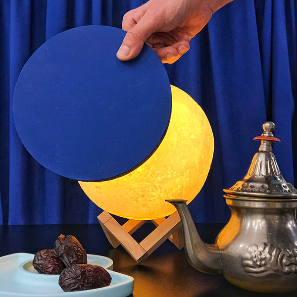 Lámpara con forma de luna y base de madera con una tetera y unos dátiles