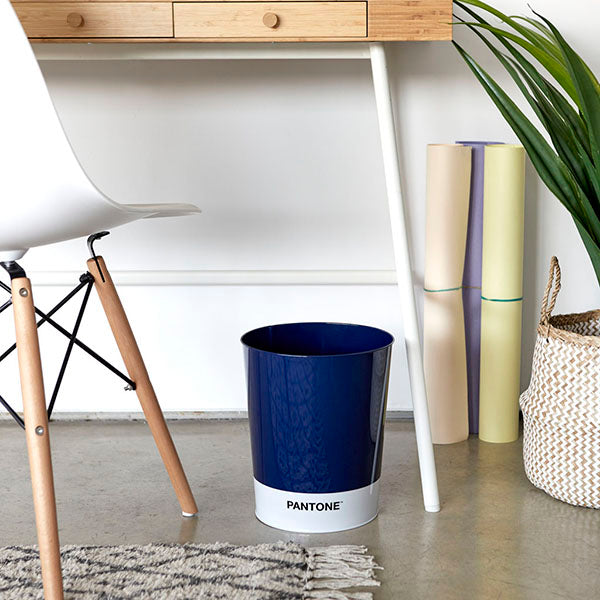 Papelera de metal azul oscuro de Pantone bajo un escritorio de madera y junto a una silla eames