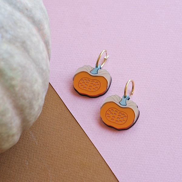 Pendientes de aro de metacrilato con forma de calabaza partida junto a una calabaza de verdad
