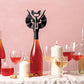 Sacacorchos de Ototo Vino con forma de murciélago negro con alas y colmillitos para usar como abridor abriendo una botella de vino rosado en una mesa puesta con velas y sobre un fondo rosa