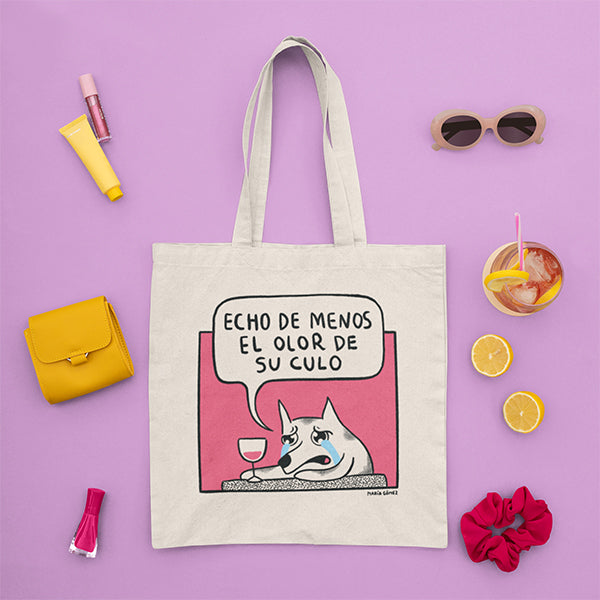 Tote bag con un perro llorando con una copa de vino mientras dice "Echo de menos el olor de su culo" rodeada de complementos